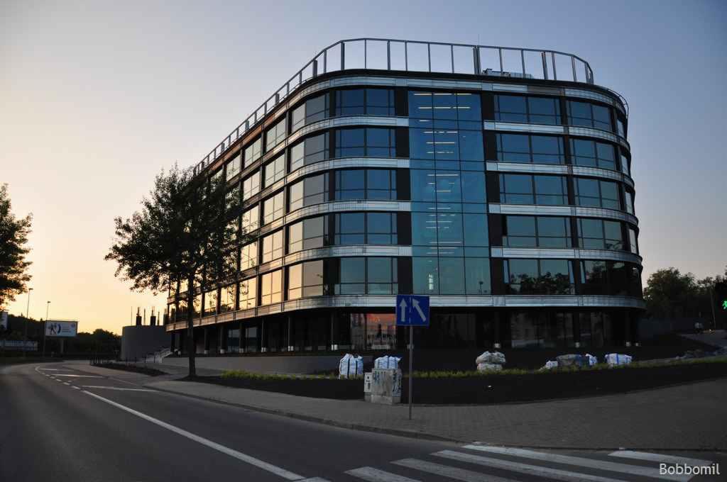 Nowoczesny budynek biurowy z zaokrąglonymi narożnikami i szklaną fasadą, widziany z poziomu ulicy o zmierzchu. W oknach odbija się zachodzące słońce, a na pierwszym planie widoczny znak drogowy.