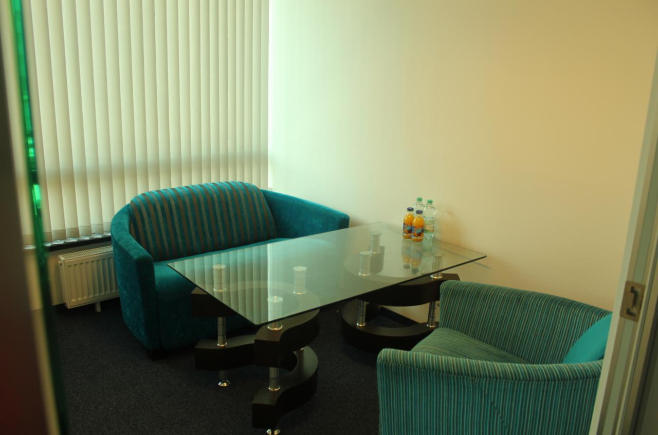 Nowoczesny pokój biurowy z turkusowymi fotelami w paski, szklanym stołem z napojami i żaluzjami w tle.