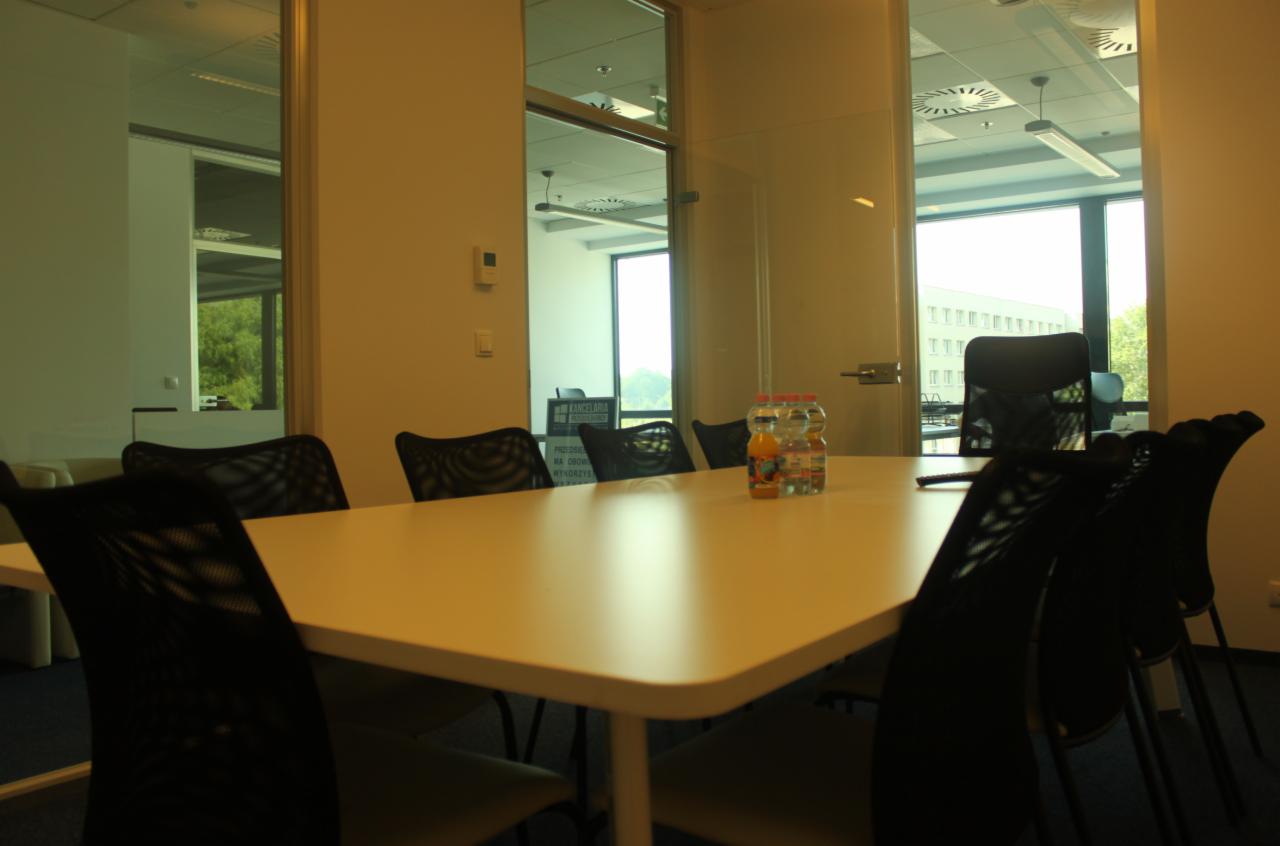 Sala konferencyjna w Szczecinie z białym stołem, czarnymi krzesłami z siatki i napojami na stole, widok przez szklane drzwi na biurowiec.