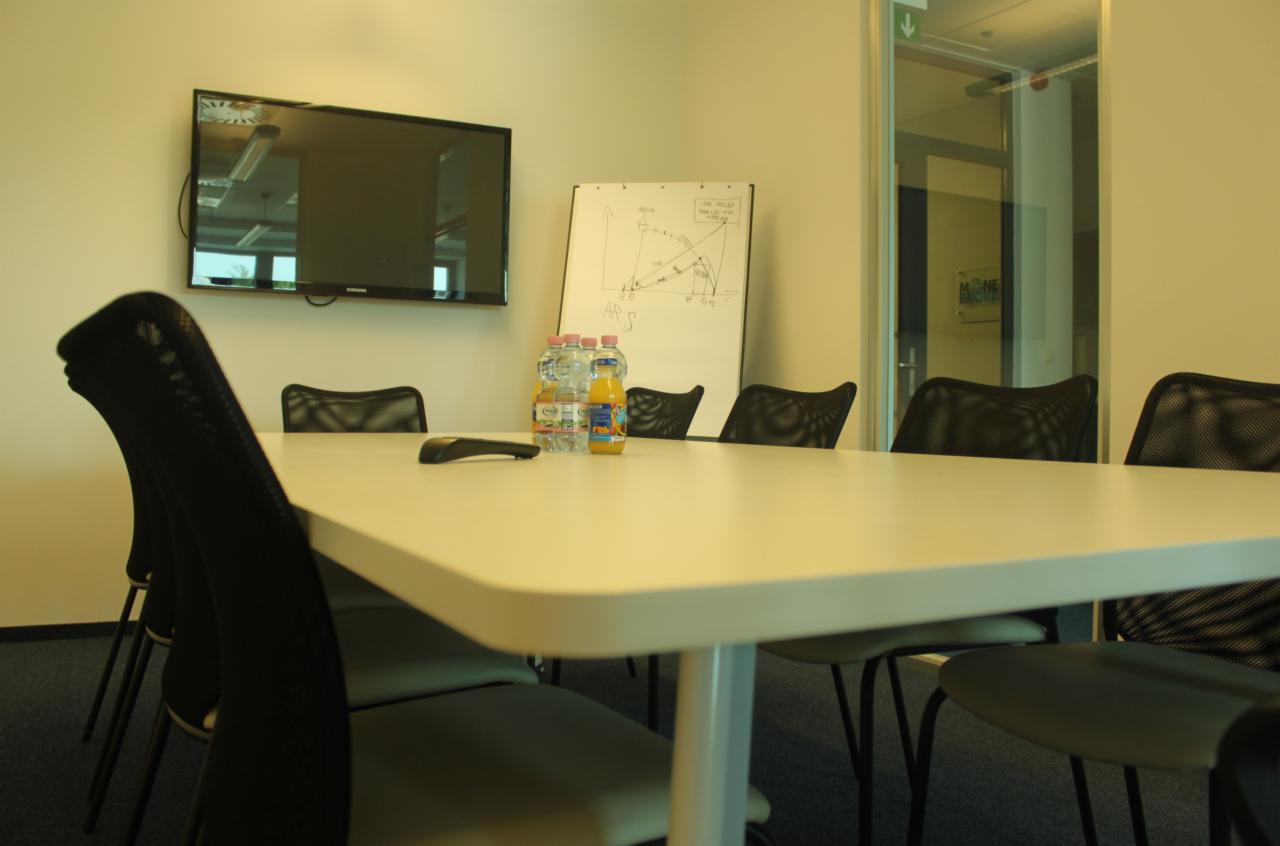 Sala konferencyjna z białym stołem, krzesłami z siatkowym oparciem, telewizorem na ścianie i flipchartem z wykresami, butelkami wody i soków na stole.