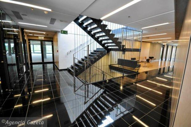 Wnętrze biurowca z czarną, błyszczącą posadzką, schodami z metalową balustradą i przeszklonymi ścianami, widoczne biurka i krzesła w oddali.