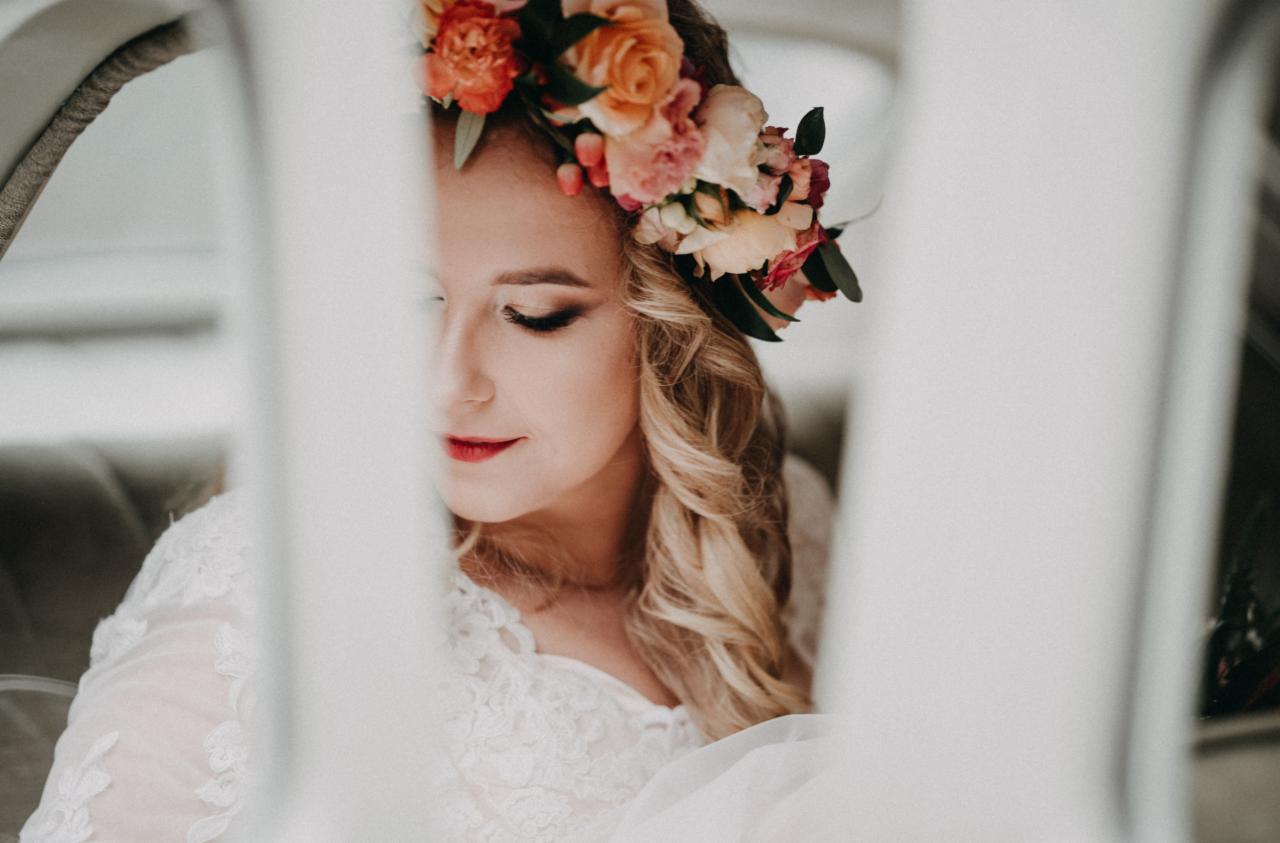 Panna młoda z kwiatowym wiankiem na głowie, delikatny makijaż, biała koronkowa suknia, ujęcie zza zasłony w samochodzie.