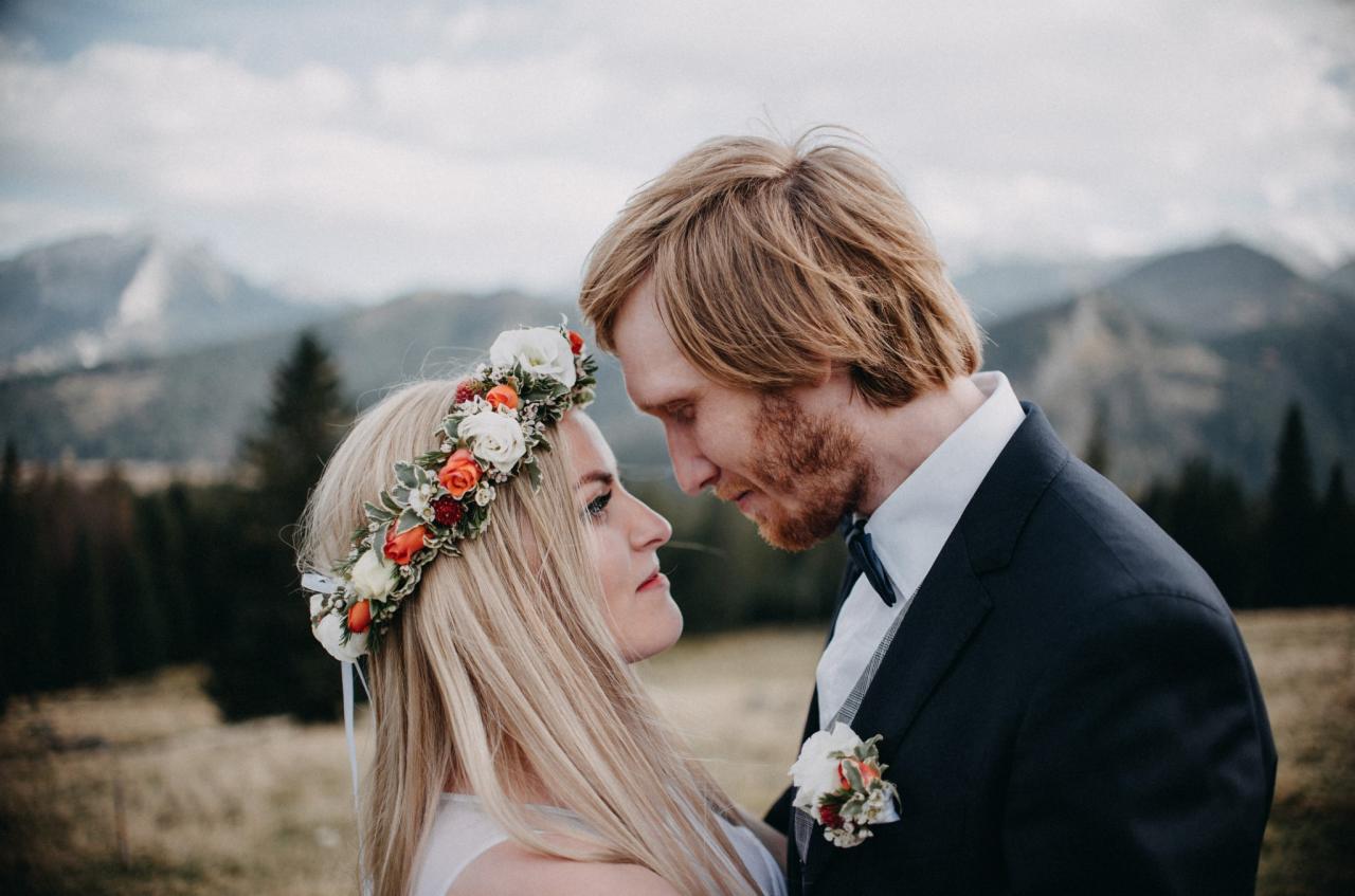 Para młoda spogląda na siebie w plenerze z górami w tle, ona w wianku z białych i pomarańczowych kwiatów, on w ciemnym garniturze z butonierką.