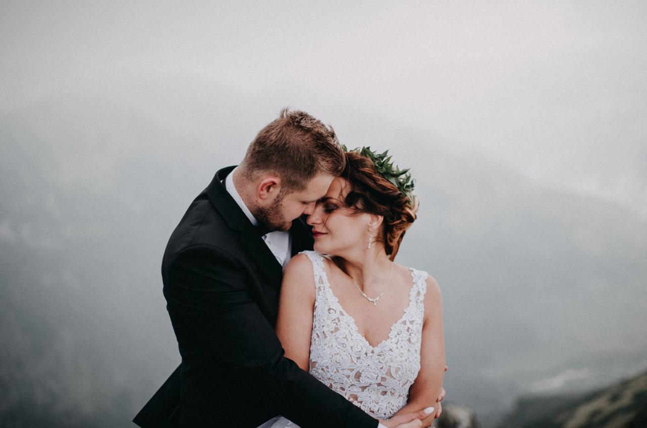 Para młoda w objęciach na tle mglistych gór, panna młoda z wiankiem z liści na włosach i koronkową sukienką, pan młody w czarnym smokingu.