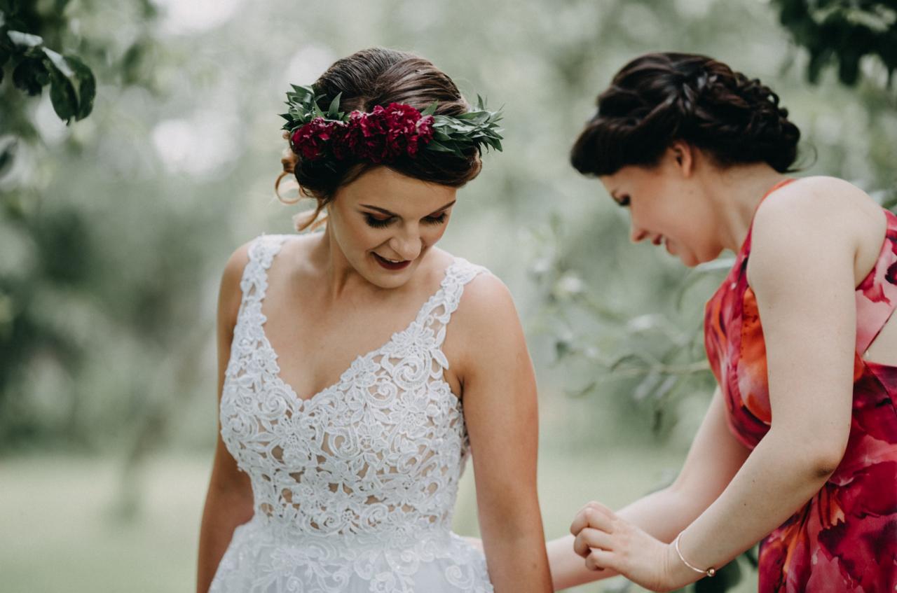 Panna młoda w koronkowej sukni z bordowymi kwiatami we włosach, poprawiana przez druhnę w kwiecistej sukience, w plenerze.