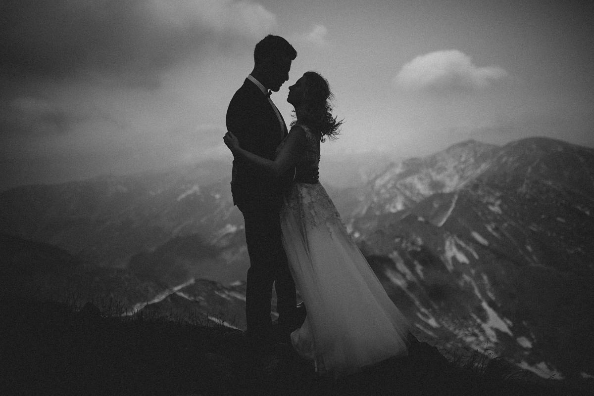 Sylwetki pary młodej w objęciach na tle górskim, czarno-biała fotografia z ziarnem, suknia ślubna z delikatnymi zdobieniami, ciemne chmury na horyzoncie.