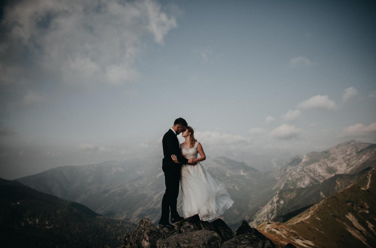 Para młoda w objęciach na szczycie skalistego wzniesienia, w tle rozległa panorama gór i pochmurne niebo.
