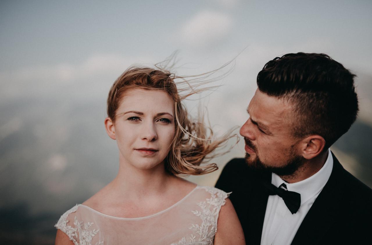 Portret narzeczeńskiej pary: kobieta w koronkowej sukni z rozwianymi włosami, mężczyzna w czarnym smokingu z muchą, z zamyślonym wyrazem twarzy na tle górskiego krajobrazu.
