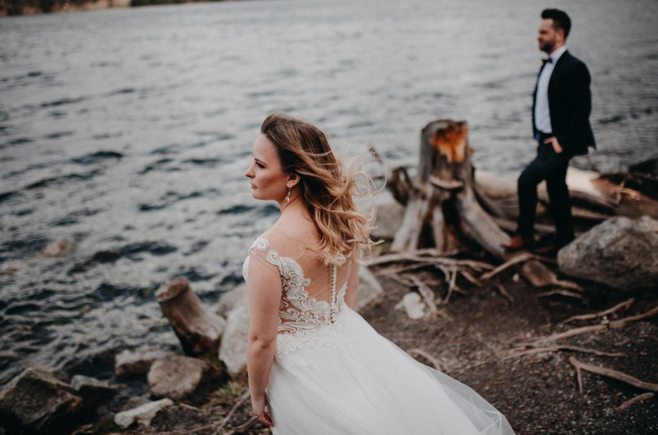 Panna młoda w zwiewnej sukni z koronkowymi zdobieniami na plecach, z rozwianymi włosami, spogląda w dal na tle jeziora w Poroninie; obok widoczny pan młody w garniturze.