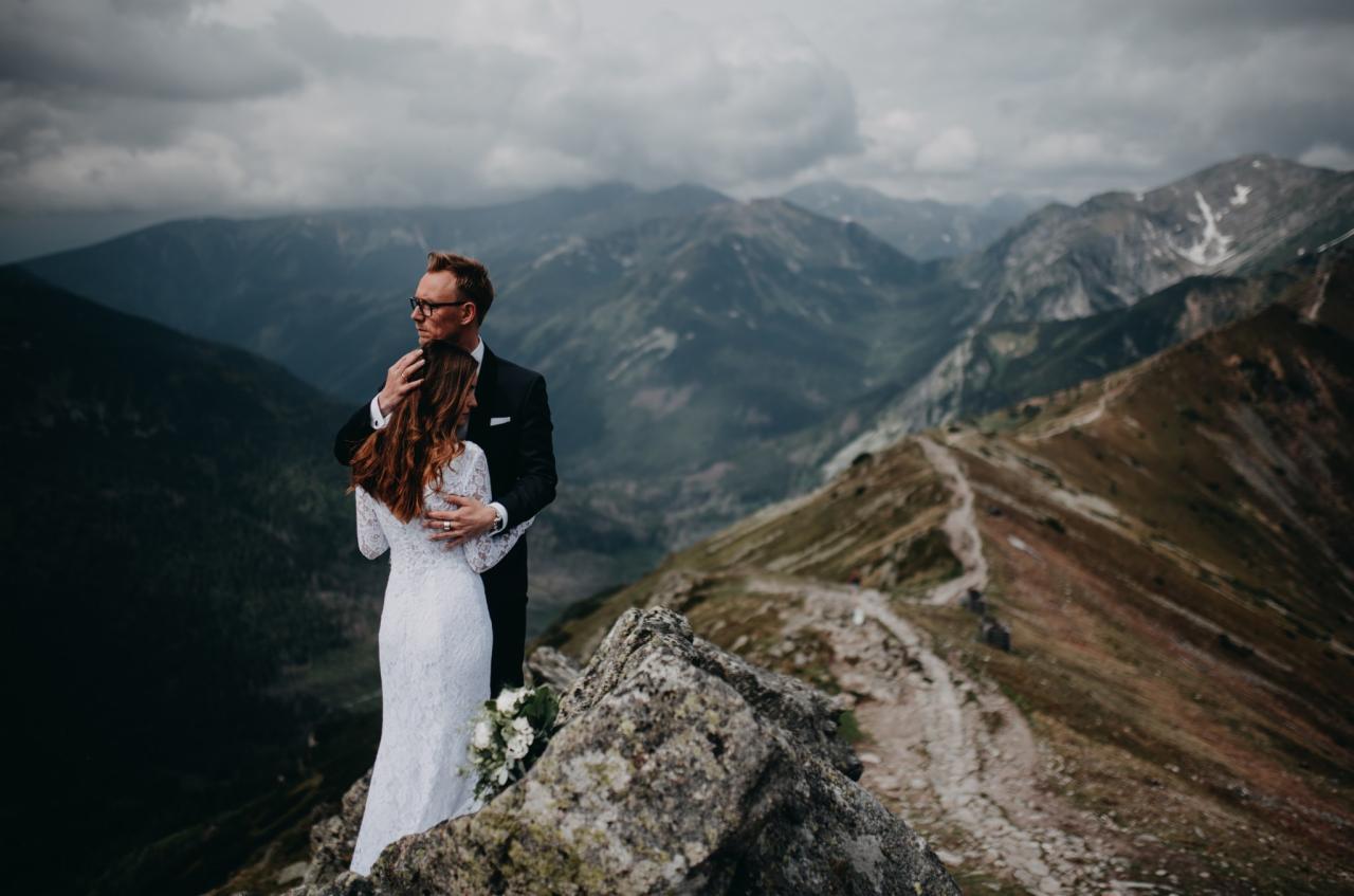 Para młoda w objęciach na szczycie góry, on w czarnym garniturze i okularach, ona w koronkowej sukni z bukietem, w tle rozległy krajobraz górski pod pochmurnym niebem.