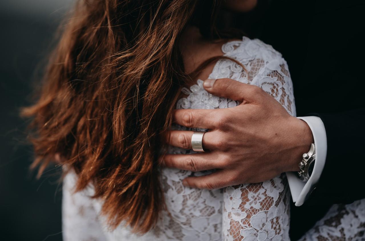 Mężczyzna obejmujący kobietę w koronkowej sukience, widoczny srebrny pierścionek i zegarek na ręce mężczyzny.