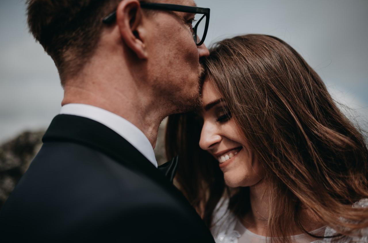 Pan młody w okularach całuje pannę młodą w czoło, ona uśmiecha się z zamkniętymi oczami.