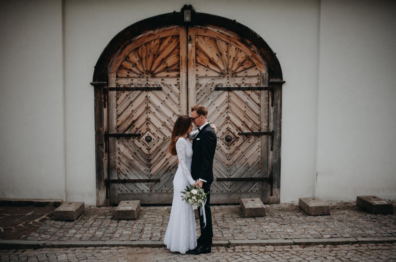 Para młoda w objęciach na tle monumentalnych, drewnianych wrót z geometrycznym wzorem, ona w koronkowej sukni z bukietem, on w klasycznym smokingu, na brukowanym placu.