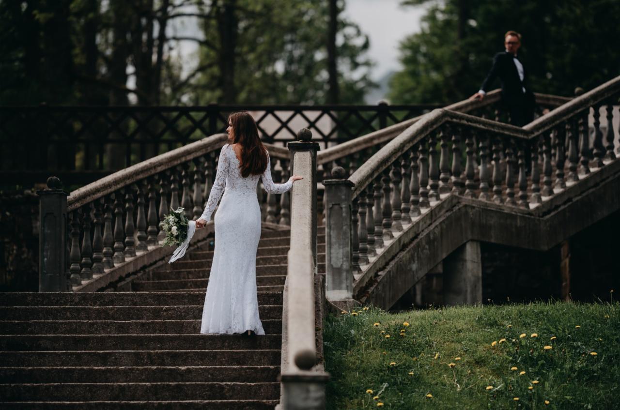 Panna młoda w koronkowej sukni wchodzi po kamiennych schodach, trzymając bukiet; pan młody czeka na górze.