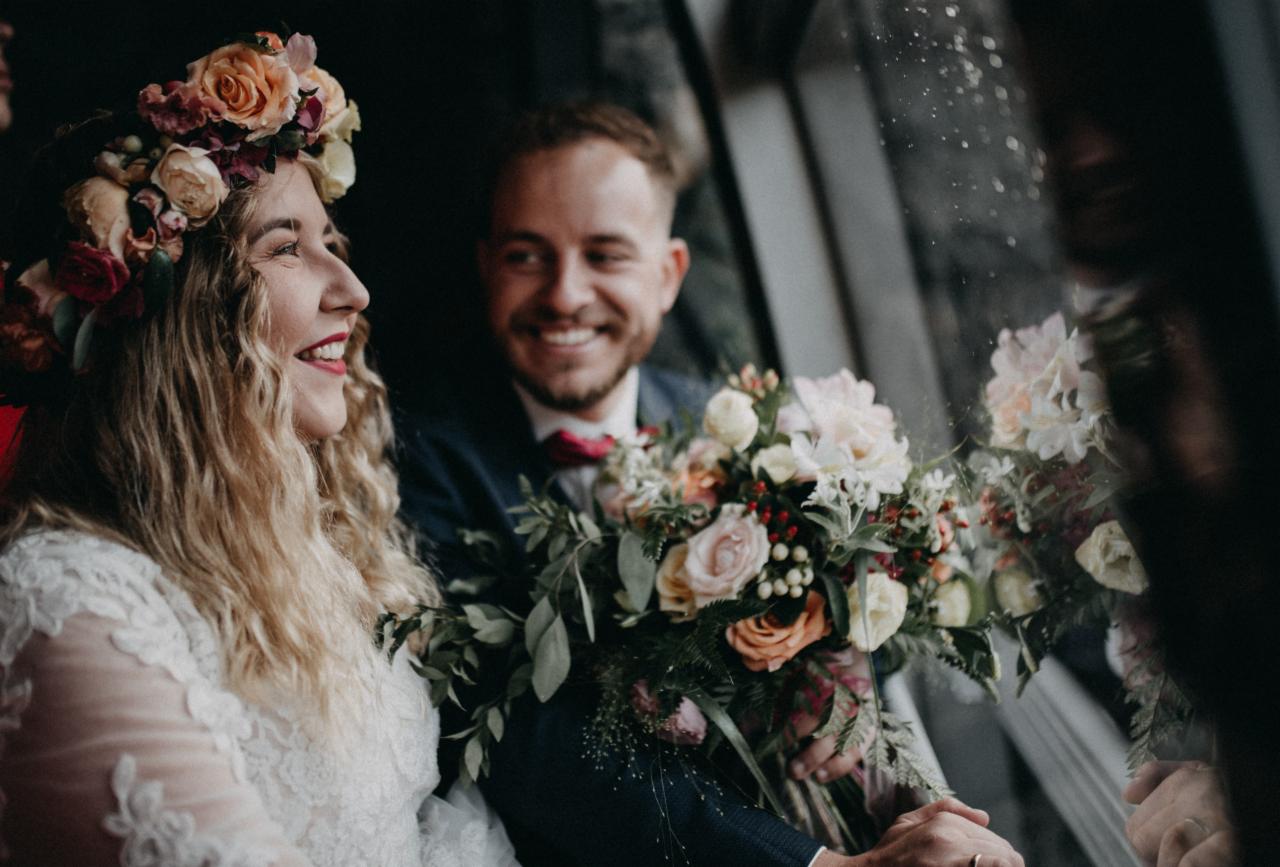 Uśmiechnięta panna młoda w wianku z kwiatów i koronkową suknią, obok uśmiechnięty pan młody w granatowym garniturze, trzymający bukiet kwiatów, widoczni w odbiciu szyby.