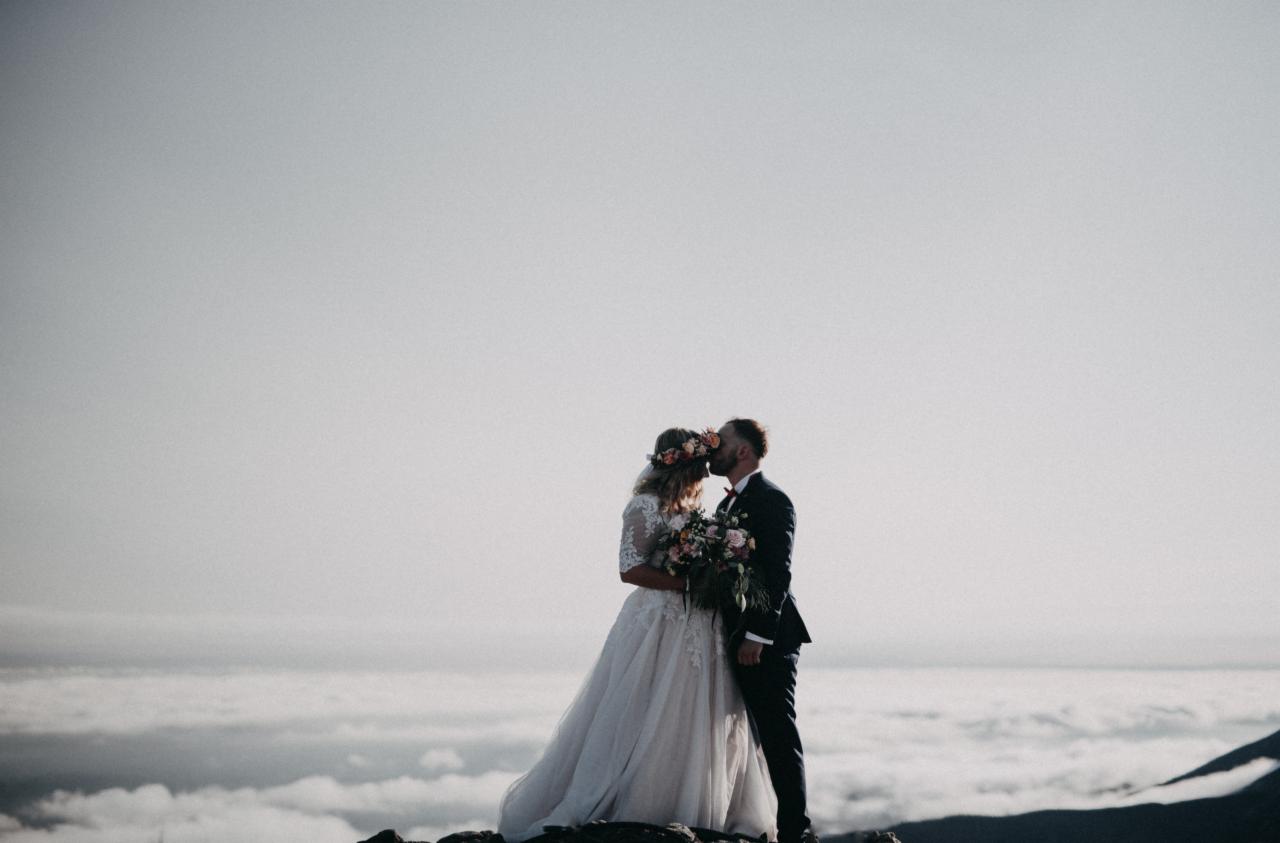 Para młoda w górach, panna młoda w koronie z kwiatów i sukni z koronkowymi rękawami, pan młody w granatowym garniturze, pocałunek na tle chmur.