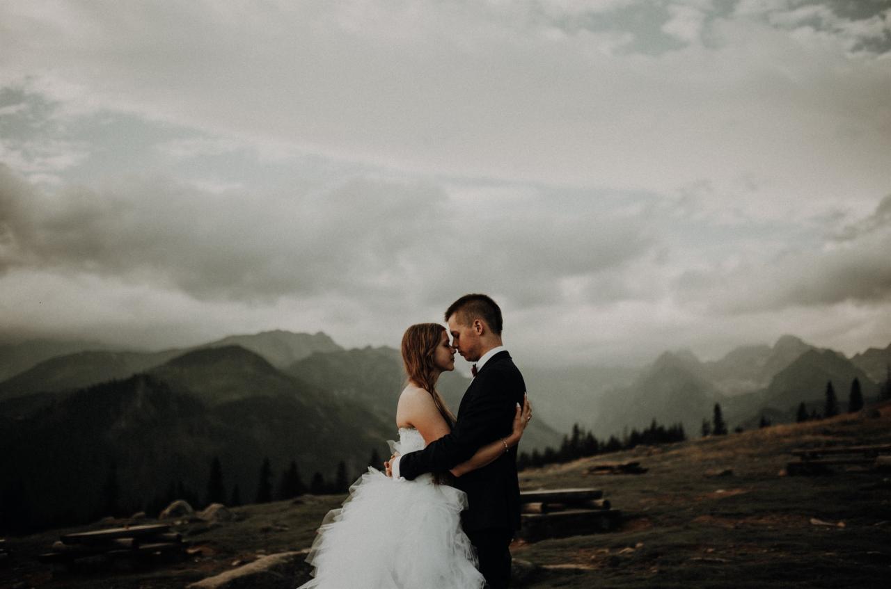 Para młoda w objęciach na tle gór i pochmurnego nieba, panna młoda w białej sukni z tiulowym dołem, pan młody w czarnym garniturze z czerwoną muchą, subtelne oświetlenie, romantyczna atmosfera.