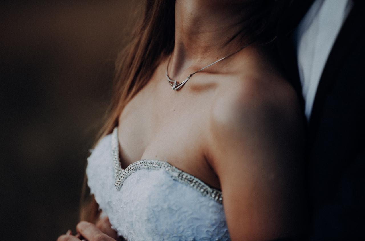 Panna młoda w białej sukni z koronkowym dekoltem i srebrnym naszyjnikiem, przytulona do mężczyzny w ciemnym garniturze.