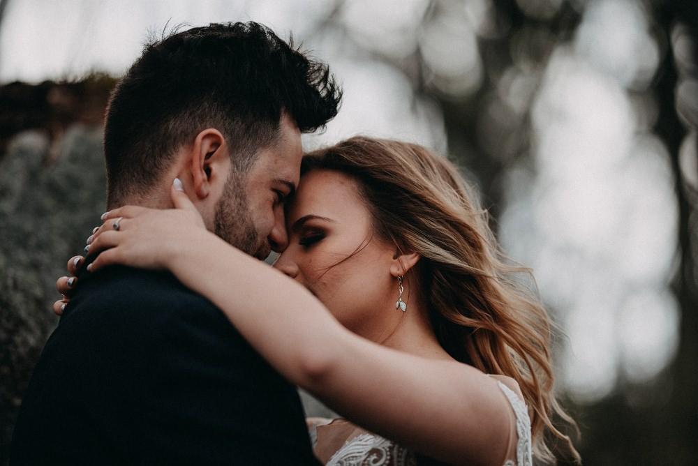 Para młoda w bliskim uścisku, panna młoda z delikatnymi kolczykami i pierścionkiem, ciemne tło z bokeh.