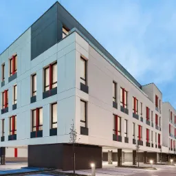 Nowoczesny budynek biurowy z białymi panelami, czerwonymi akcentami okiennymi i szarym dachem, oświetlony wieczornym światłem, widziany z perspektywy ulicy.