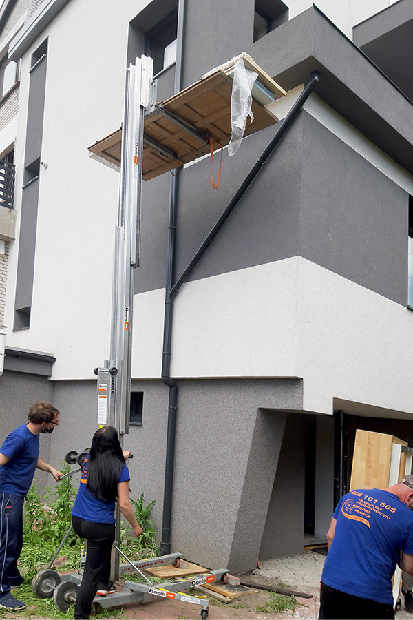 Pracownicy używają windy meblowej do wniesienia mebli na wyższe piętro budynku o szaro-białej elewacji. Na platformie windy znajdują się elementy meblowe zabezpieczone folią. Jeden z pracowników...