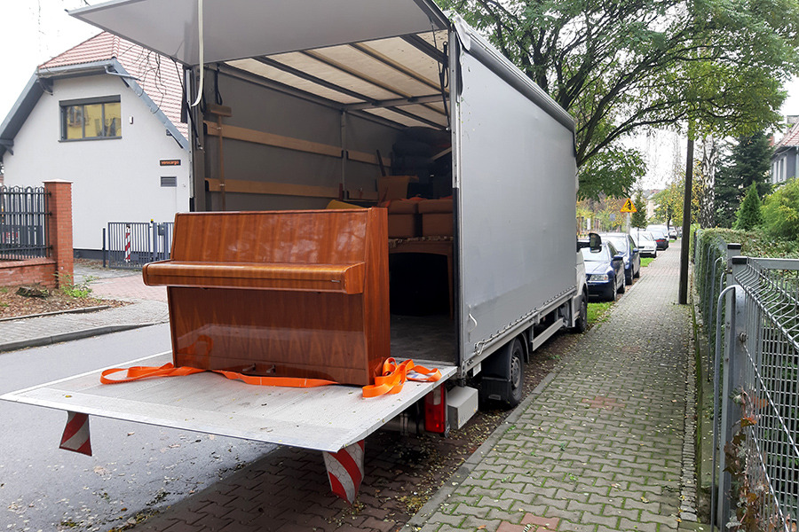 Pianino zabezpieczone pomarańczowymi pasami na platformie załadunkowej ciężarówki z otwartą przestrzenią ładunkową, w tle widoczne meble i ulica z chodnikiem z kostki.