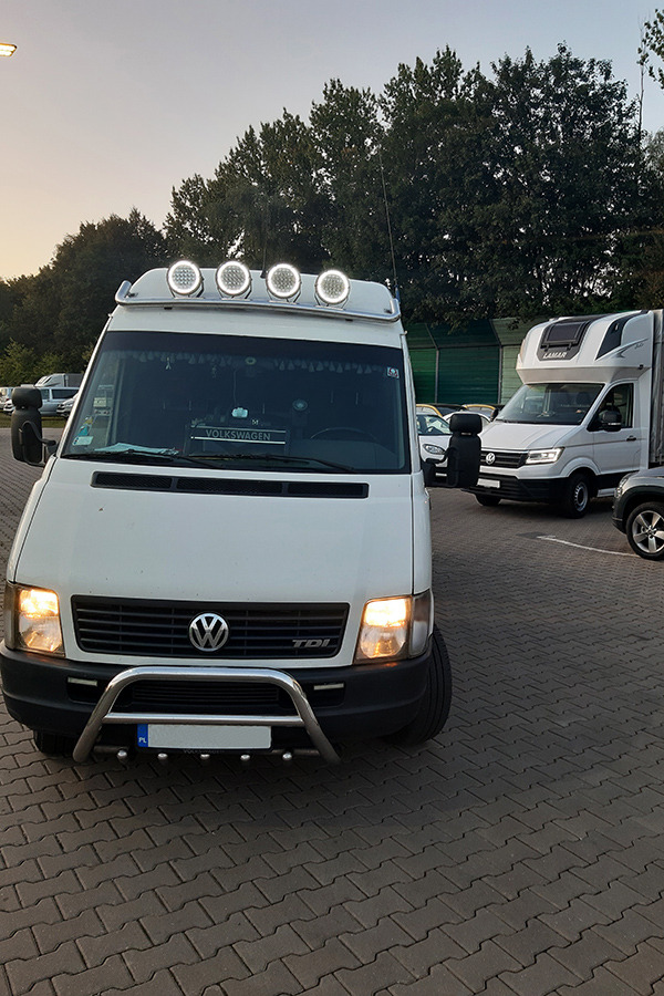 Biały Volkswagen LT z dodatkowym oświetleniem na dachu, zaparkowany w Olkuszu na kostce brukowej. W tle inne busy i drzewa.