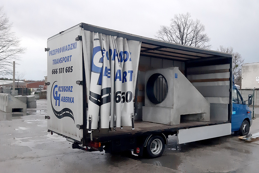 Ciężarówka z otwartą plandeką, przewożąca betonowe elementy kanalizacyjne z okrągłym otworem, na boku widoczne logo firmy transportowej z numerem telefonu.