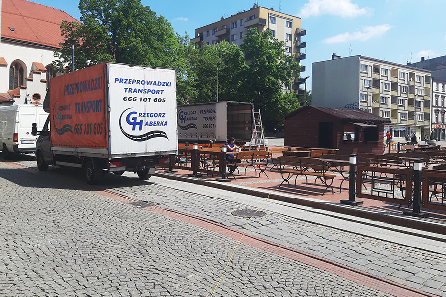 Dwa busy transportowe firmy z logotypem GH Grzegorz Aberka zaparkowane na brukowanym placu miejskim, jeden z plandeką w kolorze pomarańczowym, drugi biały z otwartymi drzwiami, w tle budynki...