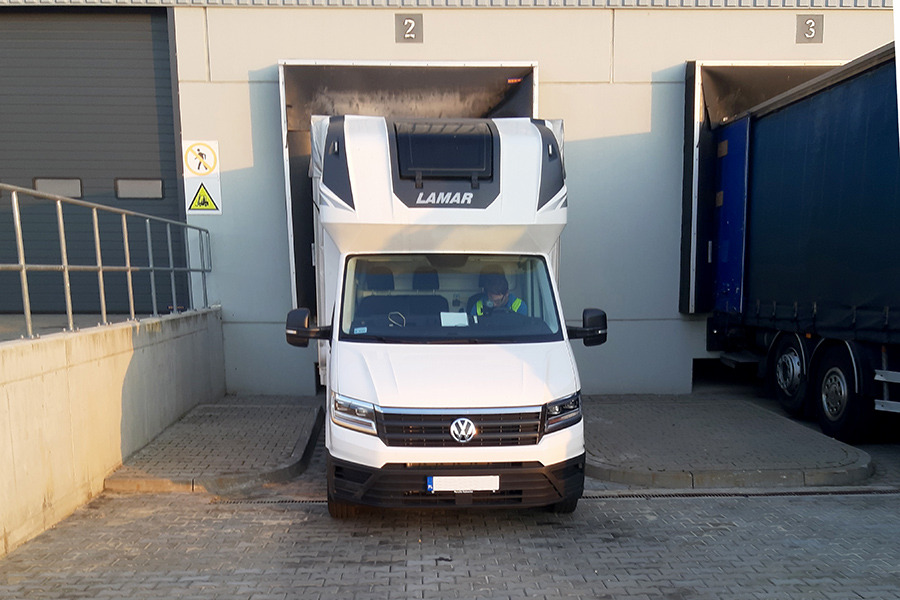 Biały bus dostawczy marki Volkswagen z nadbudową LAMAR, zaparkowany przed rampą magazynową z widocznym kierowcą w kabinie.