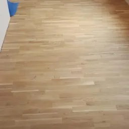 Jasna, drewniana podłoga z parkietu ułożona w pokoju, widoczna niebieska szufelka w rogu.