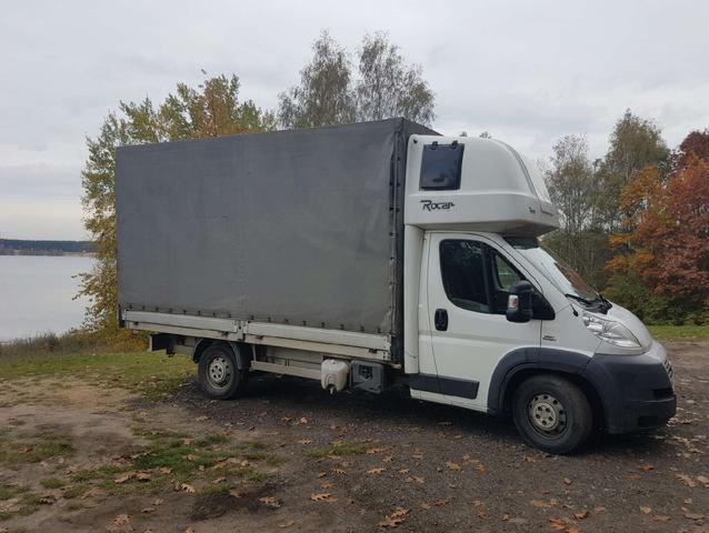 Biały samochód dostawczy z szarą plandeką na tle jeziora i jesiennych drzew, widoczny z boku, z napisem 'Rocar' na kabinie.