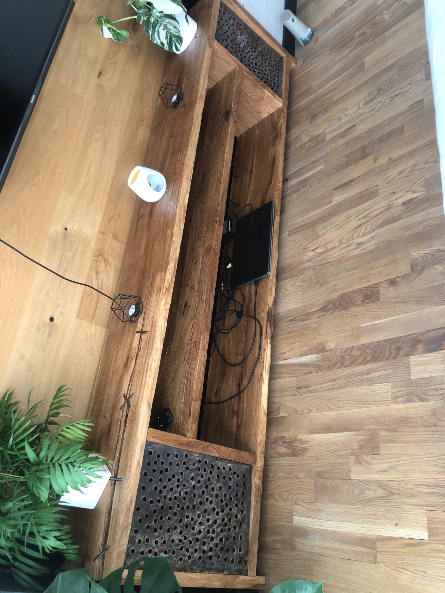 Drewniana szafka RTV z perforowanymi frontami, na parkiecie z jasnego drewna. Na szafce dekoracje: rośliny w doniczkach, geometryczne świeczniki. Widoczny fragment telewizora.