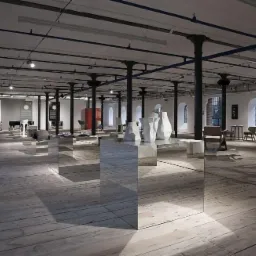 Przestronne wnętrze loftu z drewnianą podłogą, wysokimi sufitami podtrzymywanymi przez czarne, żeliwne kolumny. Na pierwszym planie stoliki z lustrzanymi bokami, na których stoją białe...