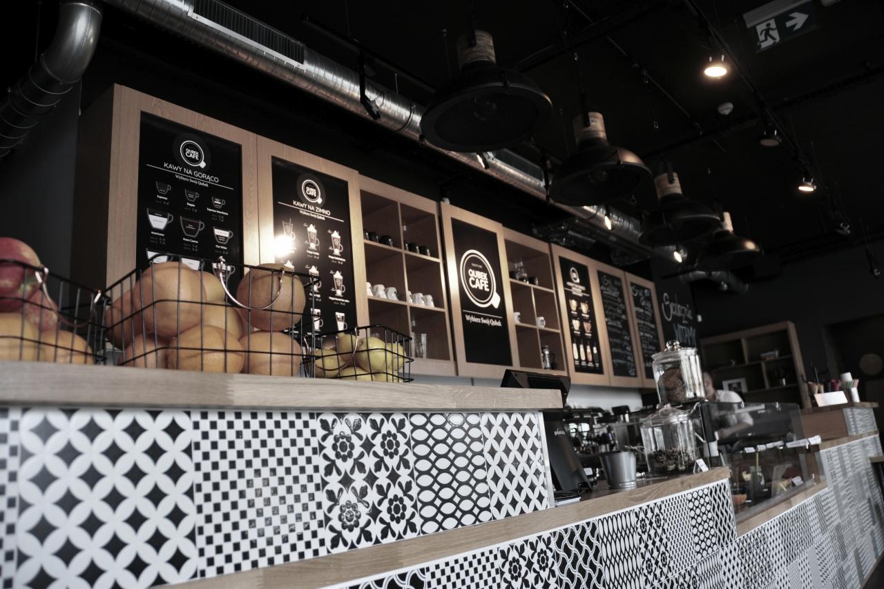 Wnętrze kawiarni z blatem wykończonym czarno-białą mozaiką, kosze z owocami na barze, menu kawowe na ścianie i wiszące lampy w stylu industrialnym.