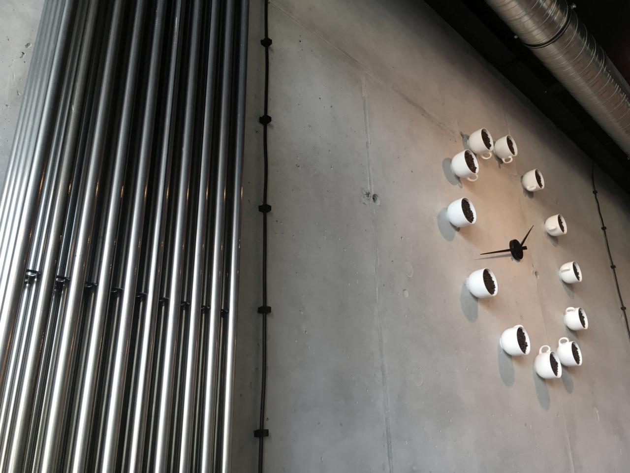 Betonowa ściana z dekoracyjnym zegarem z filiżanek kawy i metalowymi rurami biegnącymi wzdłuż ściany
