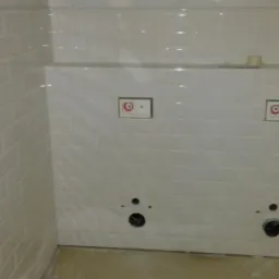 Świeżo położone, błyszczące, białe płytki typu metro na ścianach łazienki, z widocznymi otworami na przyłącza hydrauliczne i puszkami instalacyjnymi pod armaturę.