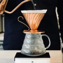 Miedziany dripper V60 z filtrem papierowym umieszczony na szklanym dzbanku, z kt&oacute;rego wylewa się woda z miedzianego czajnika o długiej wylewce, całość stoi na wadze.