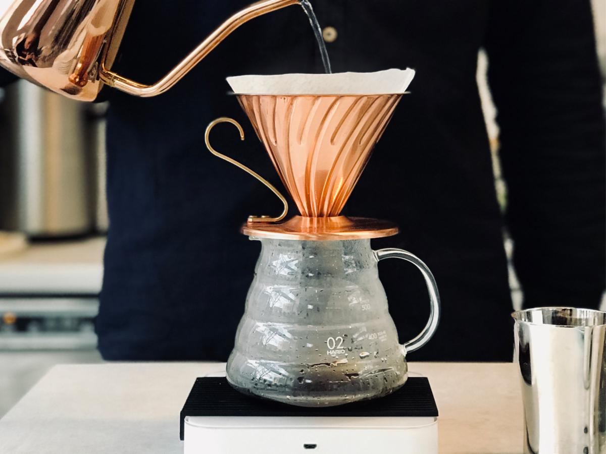 Miedziany dripper V60 z filtrem papierowym umieszczony na szklanym dzbanku, z którego wylewa się woda z miedzianego czajnika o długiej wylewce, całość stoi na wadze.