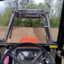 Widok z kabiny traktora z ładowaczem czołowym wyrównującego teren pod trawnik w Rucianem-Nidzie, widoczna świeżo przygotowana gleba i drzewa w tle.