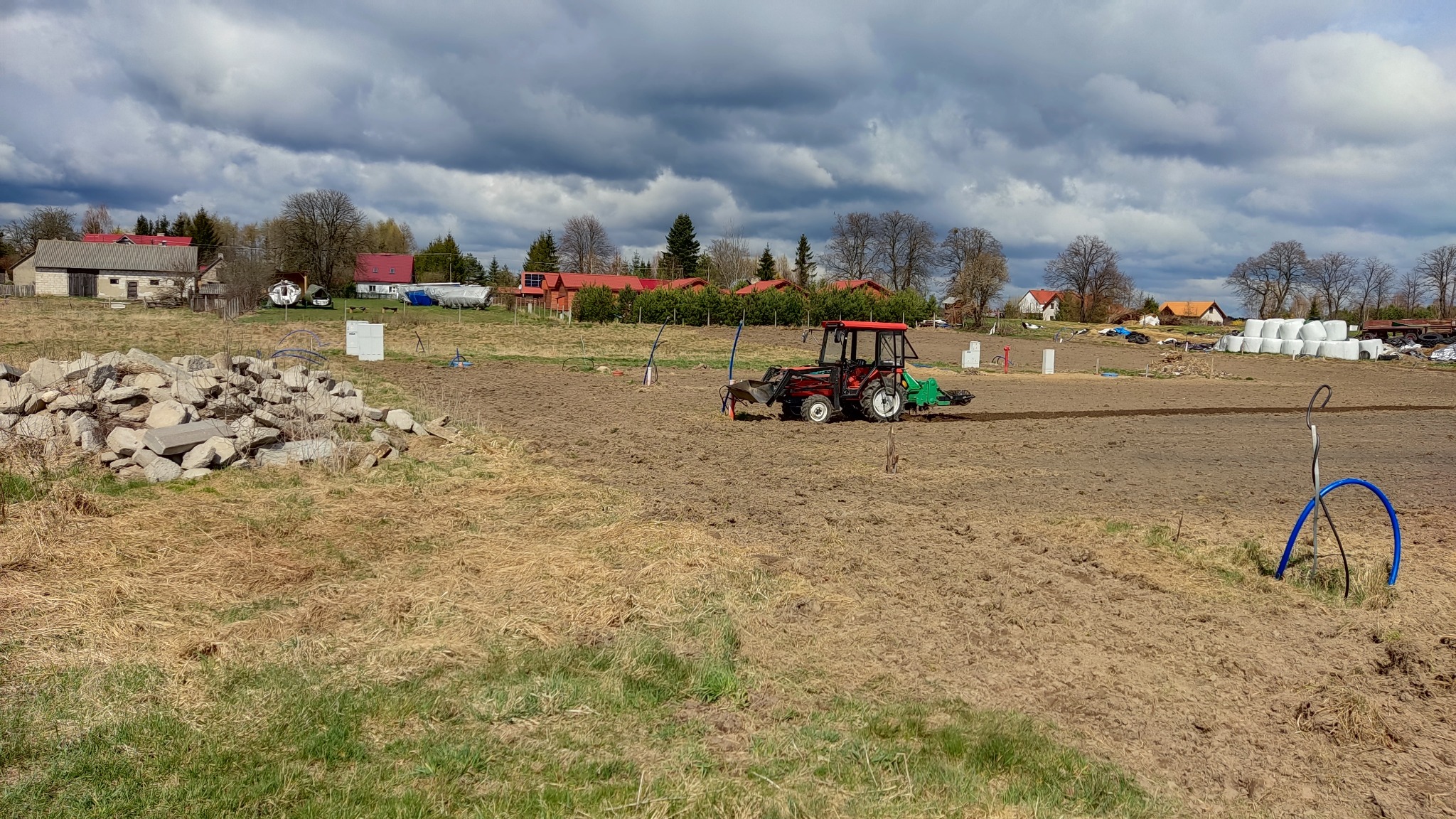 Czerwony traktor z glebogryzarką przygotowuje pole pod uprawę, w tle sterta kamieni, budynki gospodarcze i pochmurne niebo.
