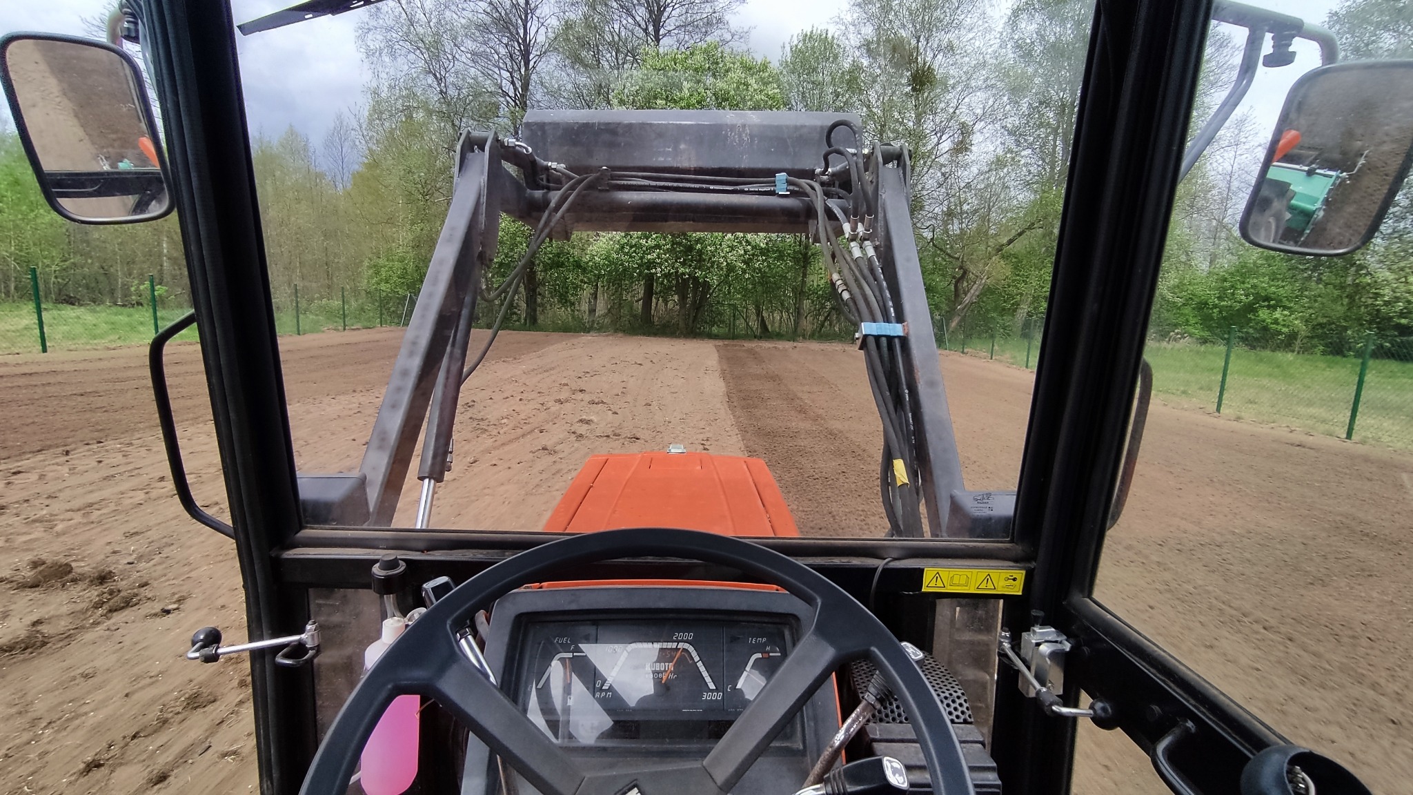 Widok z kabiny traktora z ładowaczem czołowym wyrównującego teren pod trawnik w Rucianem-Nidzie, widoczna świeżo przygotowana gleba i drzewa w tle.