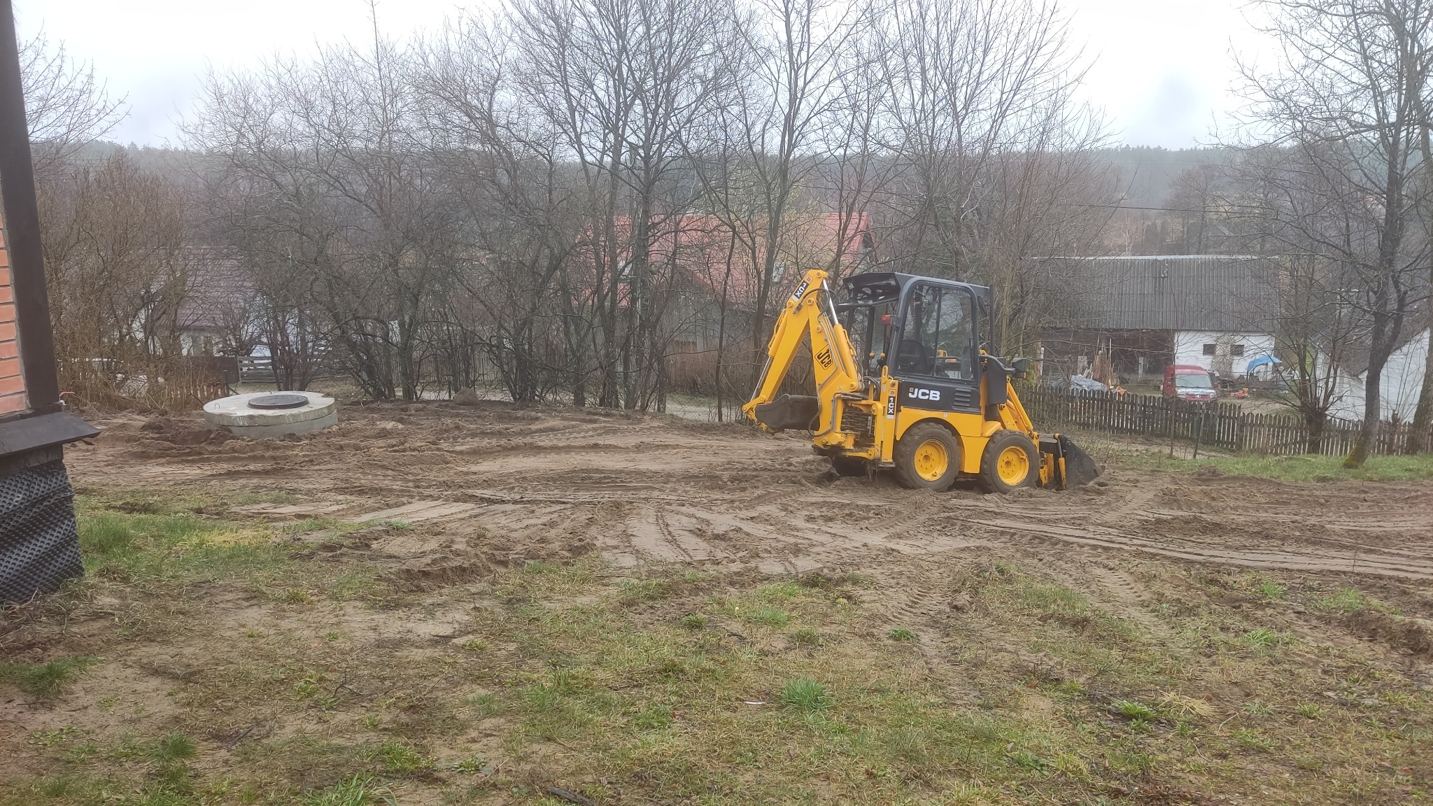 Żółta mini koparka JCB na błotnistym placu budowy, w tle widoczny betonowy krąg studni i zabudowania wiejskie w pochmurny dzień.