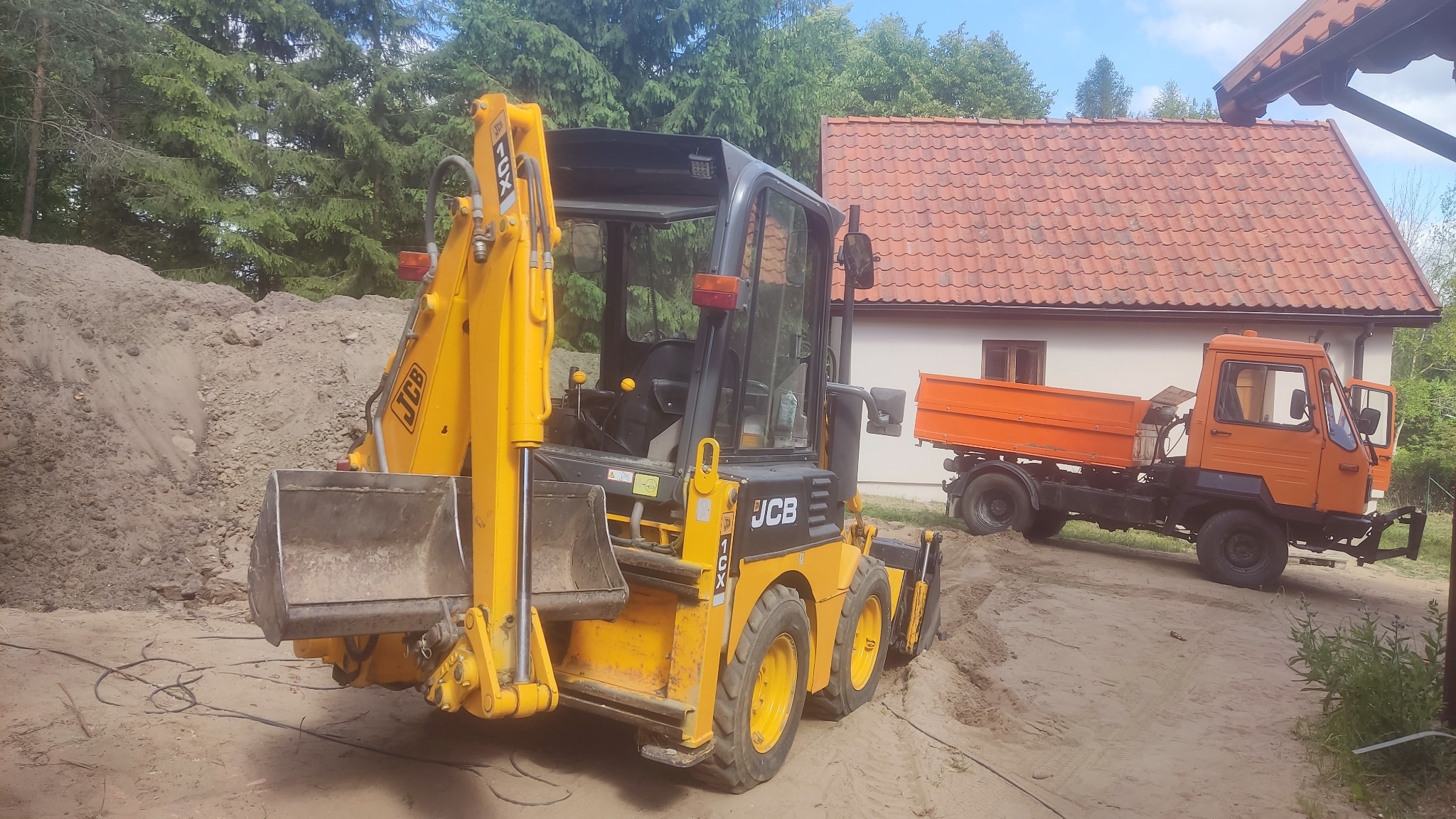 Żółto-czarna koparko-ładowarka JCB z łyżką z przodu, obok pomarańczowa wywrotka, w tle dom z czerwoną dachówką i hałda ziemi.