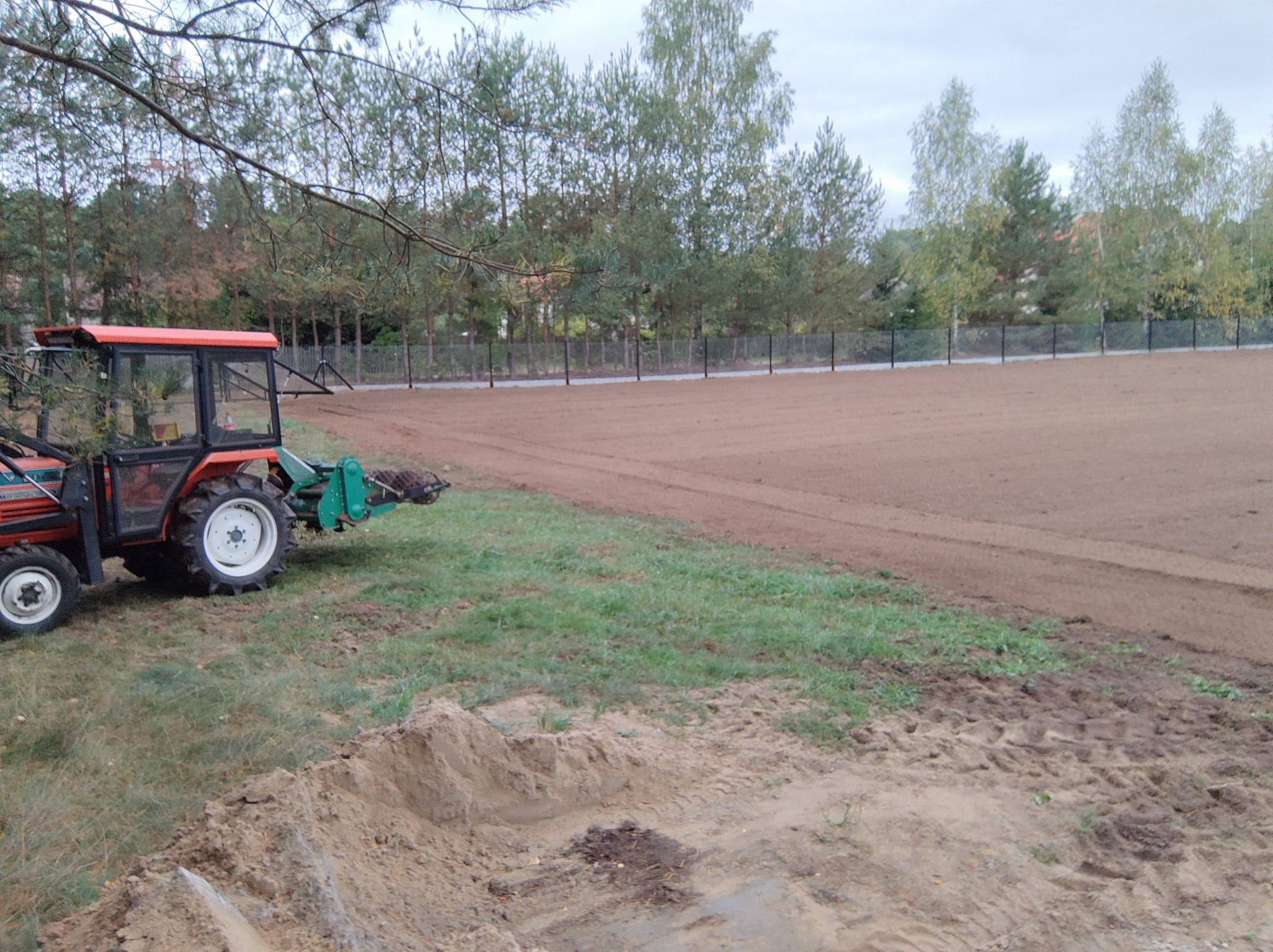 Traktor z glebogryzarką przygotowuje teren pod trawnik; widoczny świeżo wyrównany obszar, obok pryzma ziemi i trawa, w tle drzewa i ogrodzenie.