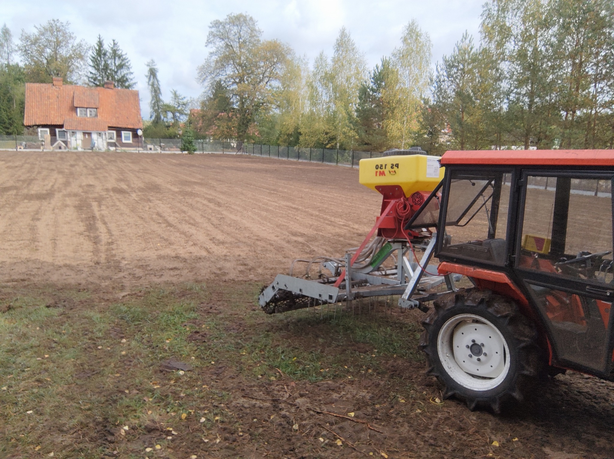 Traktor z siewnikiem na przygotowanym polu, w tle dom z czerwoną dachówką i drzewa.