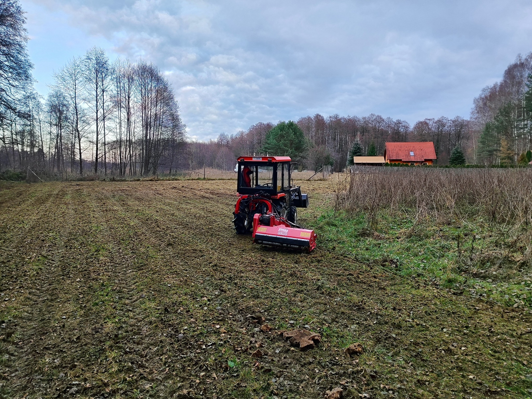 Czerwony traktor z glebogryzarką na polu, przygotowanym pod uprawę, w tle widoczny dom z czerwoną dachówką i drzewa bez liści pod pochmurnym niebem.