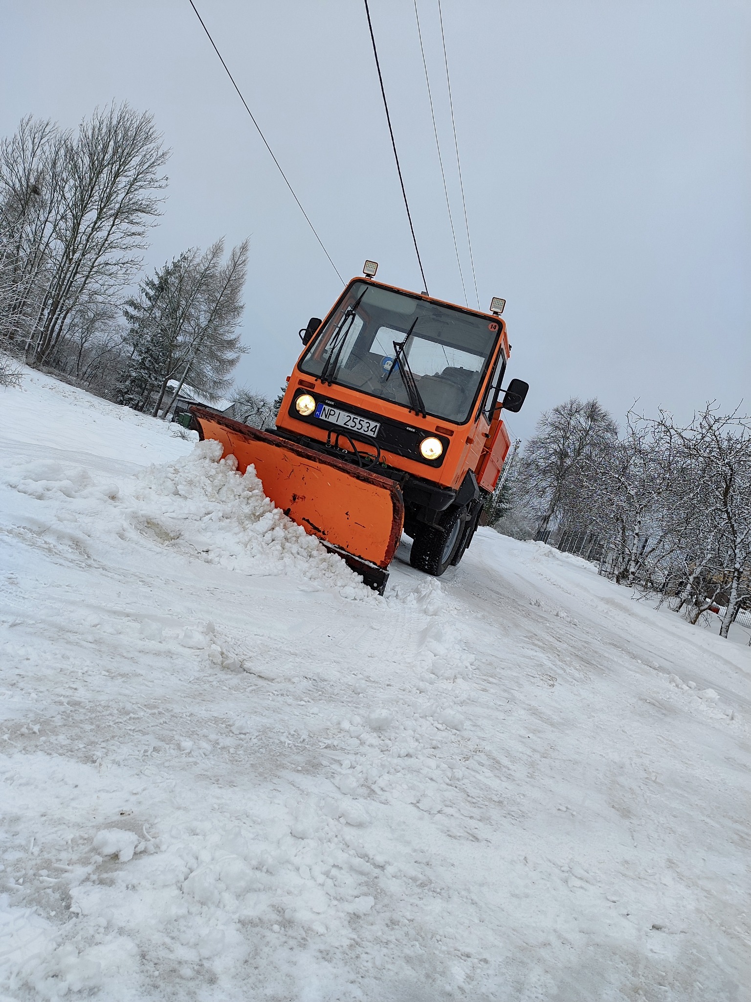 Pomarańczowy pług śnieżny usuwa śnieg z zasypanej drogi w zimowy dzień, widoczne drzewa pokryte śniegiem w tle.
