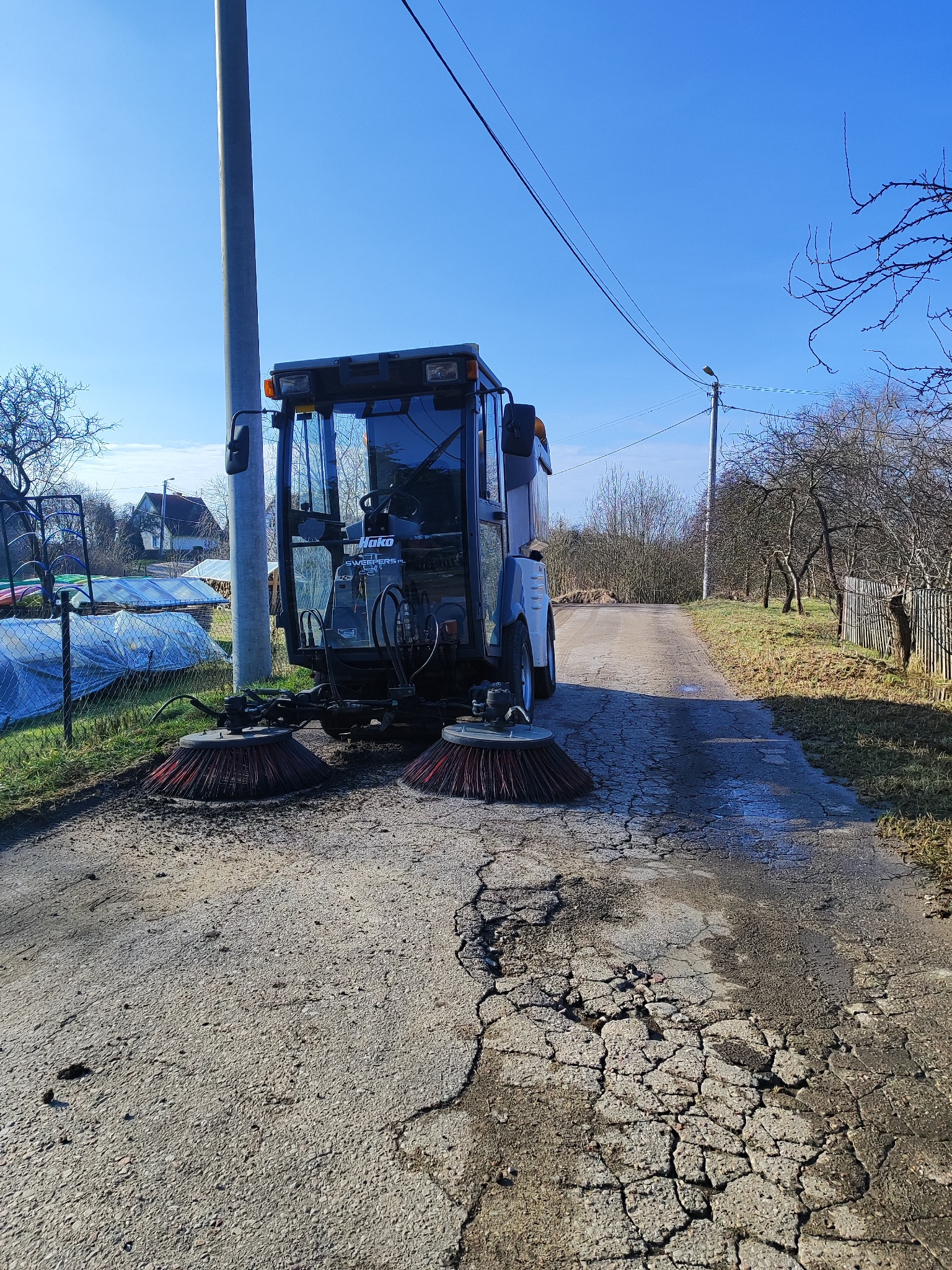 Maszyna czyszcząca Hako na zniszczonej asfaltowej drodze, z widocznymi szczotkami i otoczeniem wiejskim.