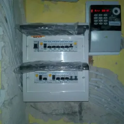 Nowa skrzynka bezpiecznikowa marki NEXT, jeszcze w folii ochronnej, zamontowana obok licznika energii elektrycznej na surowej ścianie w trakcie prac remontowych.